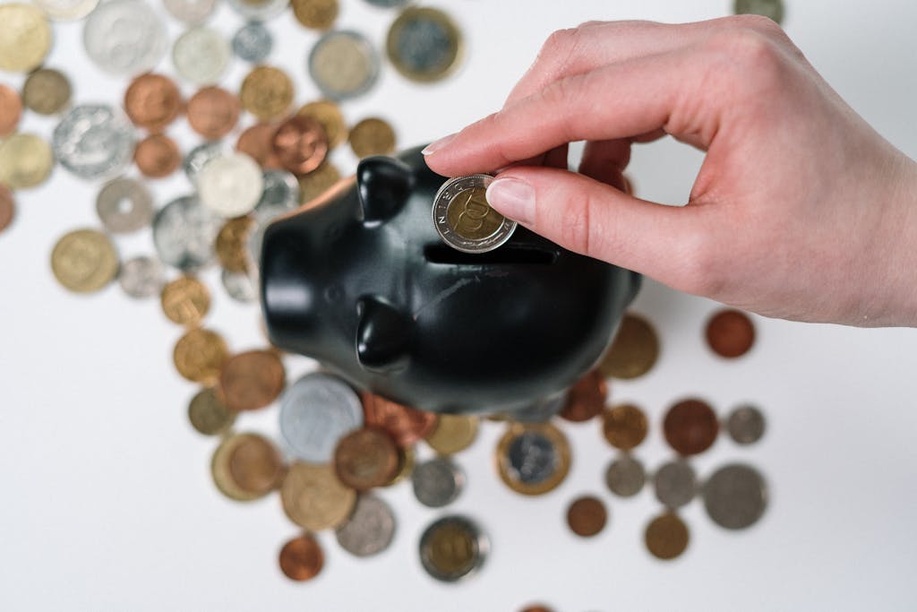 Close-up of a hand inserting a coin into a black piggy bank with scattered coins on a white background.